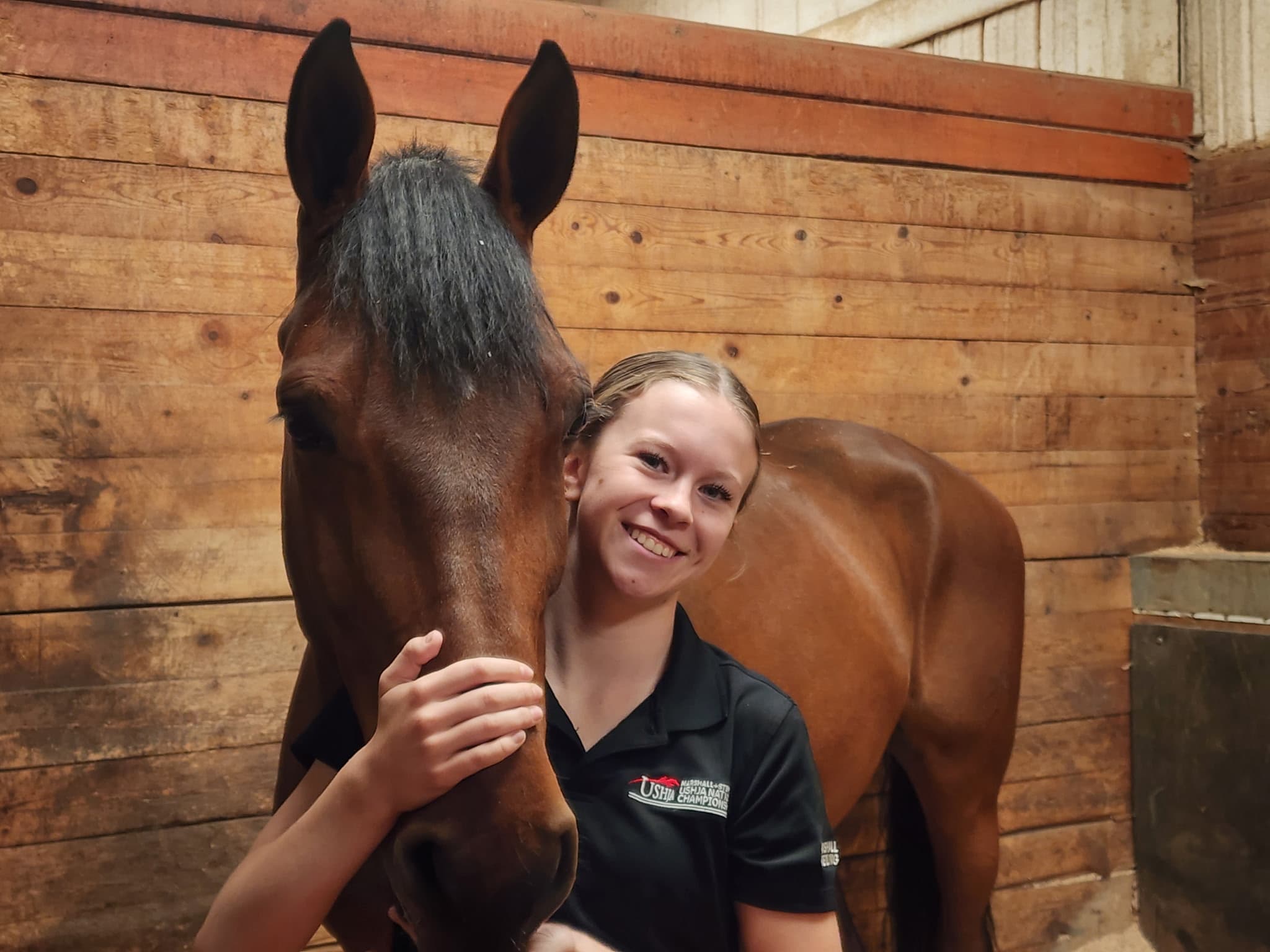 Tully smiling next to a bay horse's head in the barn aisle.